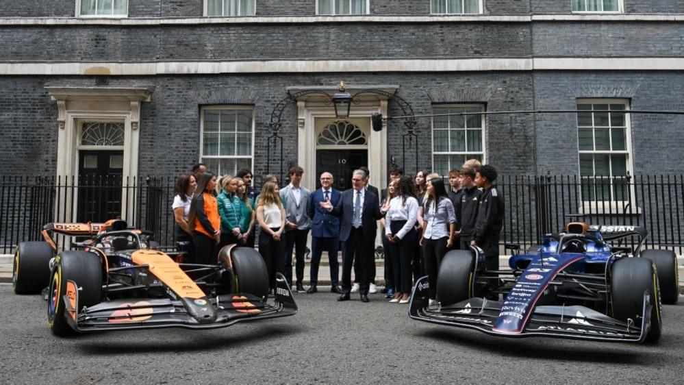 Two Formula 1 cars either side of the prime minster and several people looking at him and listening. The black door and grey brick of Number 10 Downing street is behind them