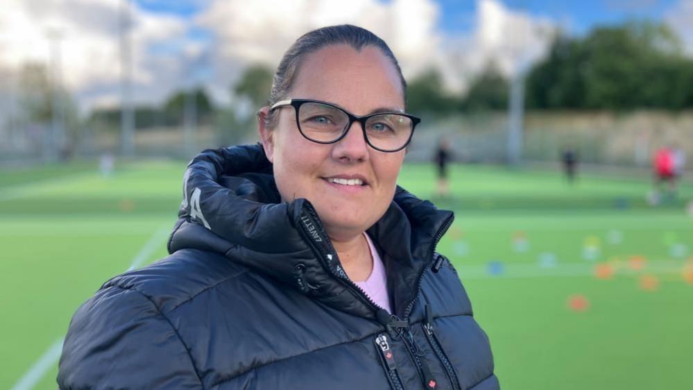 A middle-aged white woman with dark hair and glasses is standing on an artificial sports pitch and smiling. Her hair is tied back and she is wearing a padded black jacket with a high collar and hood. 