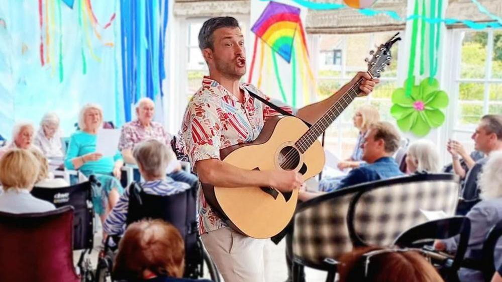 A young man holding a guitar and smiling whilst elderly people in wheelchairs look on.