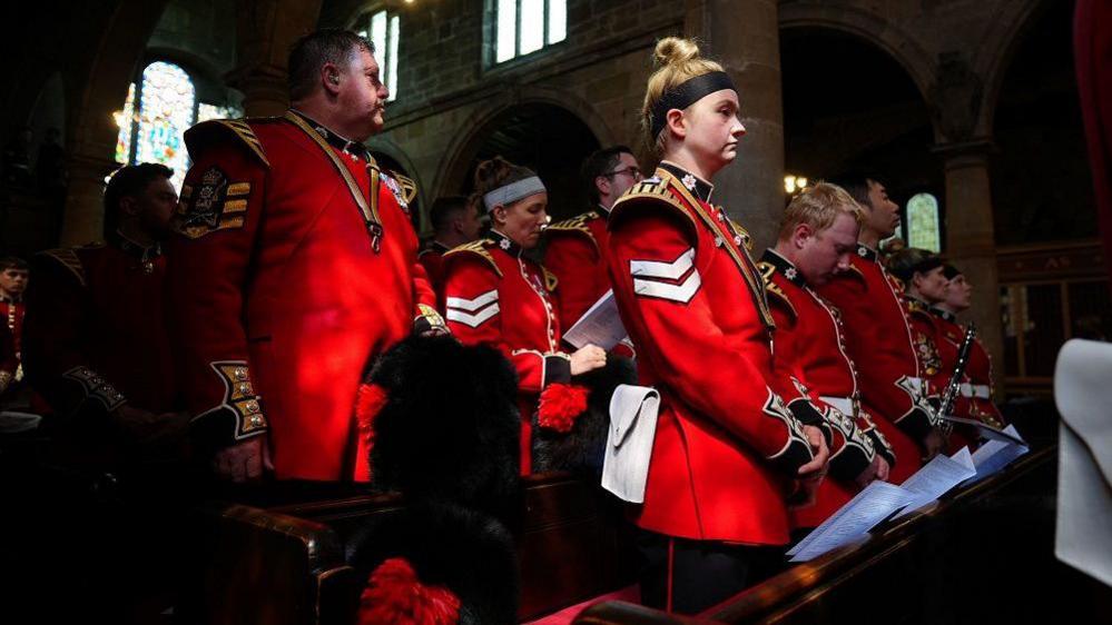 Guardsmen standing in church pews. The woman in the front of the picture has blonde hair tied up in a bun and a black headband on her head and is looking straight ahead. The guards around her are reading from sheets of paper.