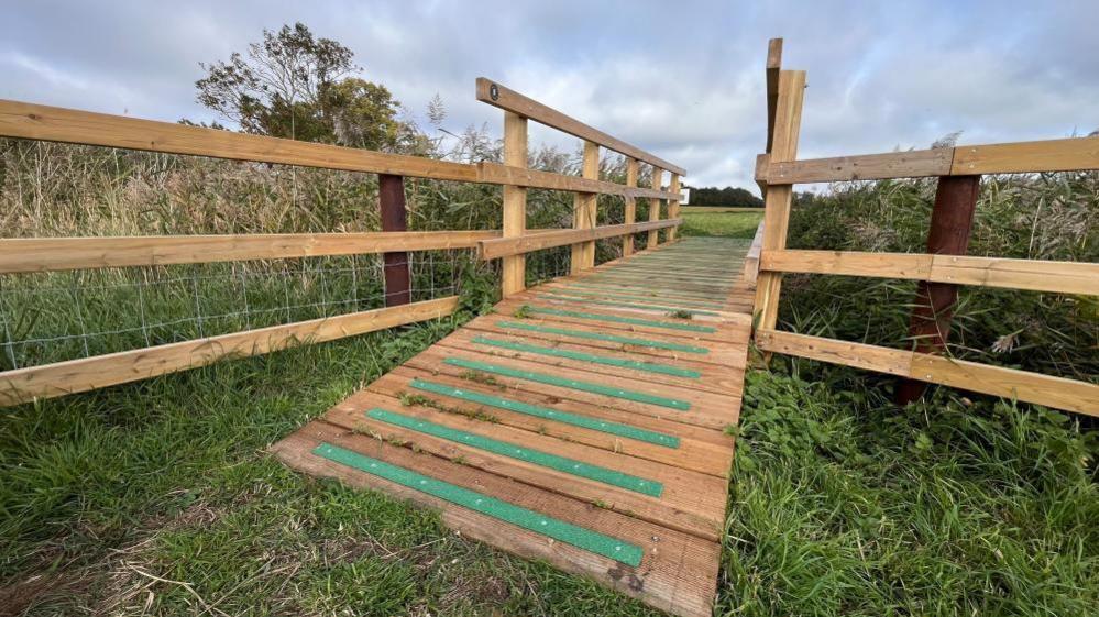 There is a small wooden bridge over a ditch. It has a ramp with green strips for grip and new fencing.