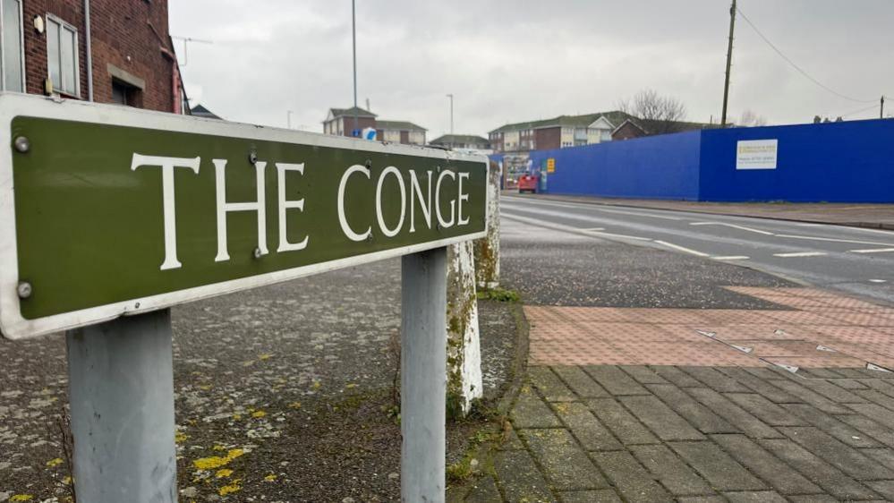 The Conge, shown on a road sign, with tactile paving for a pedestrian crossing visible, and blue hoarding screening off the development site in the right distance. Behind the roadsign is a building built of red brick.