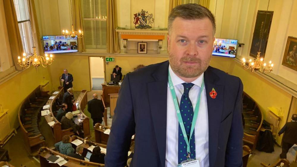 A man standing in front of a council chamber. He is wearing a tie and looking serious.