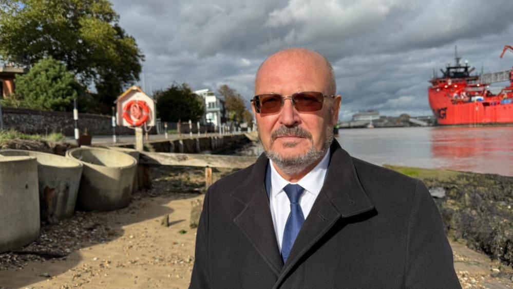 Graham Plant stands in a white shirt, blue tie and black overcoat, on the beach of the River Yare in Gorleston. He is wearing tinted prescription spectacles. On the right is an orange-painted ship, and on the left pipes and other structures shoring up Riverside Road. A life ring can be seen in a white housing, and trees and properties beyond.