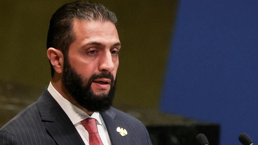 Al-Sharaa speaks at the UN wearing a pinstripe suit and red tie. 
