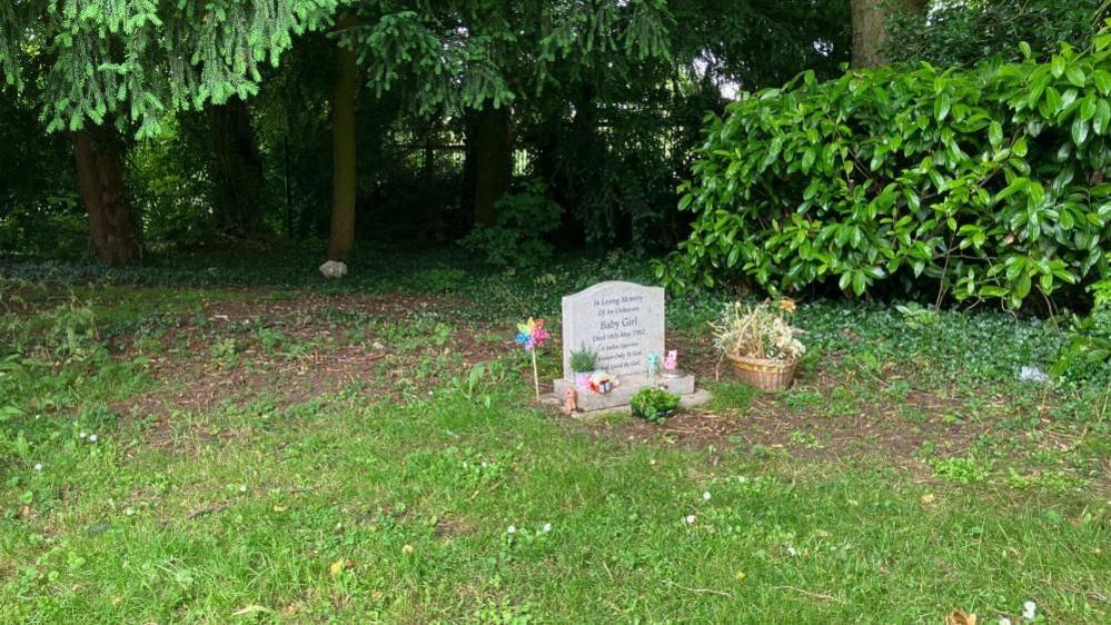 Headstone with flowers and a toy windmill set in grass with trees behind