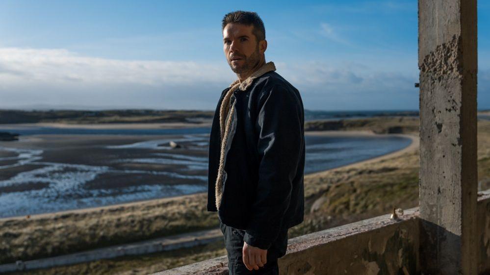 Dónall Ó Héalai  in a denim jacket and jeans, pictured in front of a Donegal bay