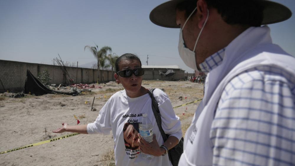A woman wearing a T-shirt with the image of a missing relative stands behind a crime-scene tape at the Izaguirre Ranch and gestures towards the sand behind here while speaking to a man.