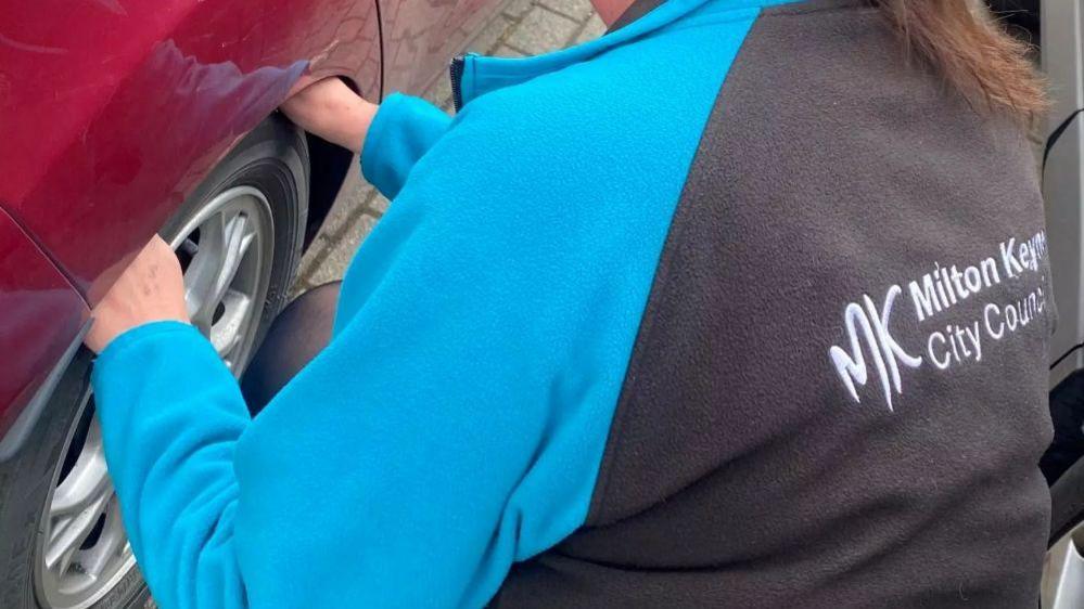 A woman, in a Milton Keynes City Council fleece, with blue sleeves and black back, holding onto a tyre on a red car. You can only see her back, and part of a ponytail.