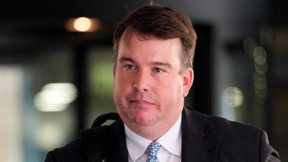 Craig Williams is walking in front of the court building. He has short brown hair and is wearing a black suit jacket, white shirt and a patterned tie.