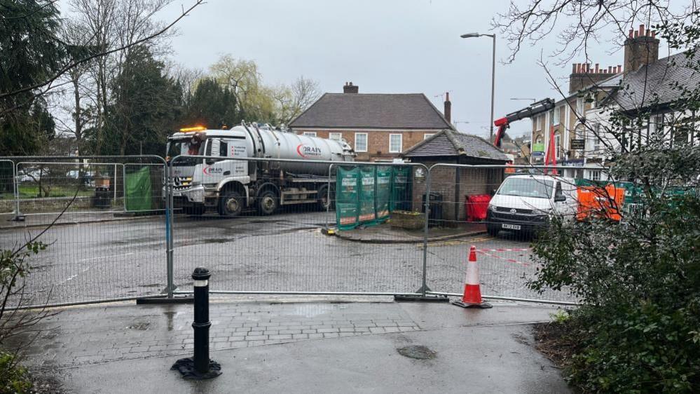 A tanker clearing water in Chalfont St Peter. It shows a road cordoned off by a metal fence, a tanker in the distance and a cone.