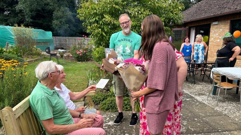 A man in a green polo and a woman in a white polo sit on a bench in a garden and are handed several bouquets of flowers 