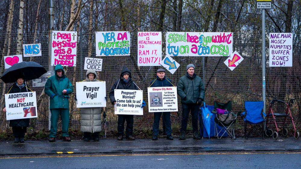 People gather by a fence holding signs saying things like "abortion is not healthcare" and "pregnant? worried? come talk to us! we can help you". Signs pinned to the fence behind them read things like "yer da protests abortion"