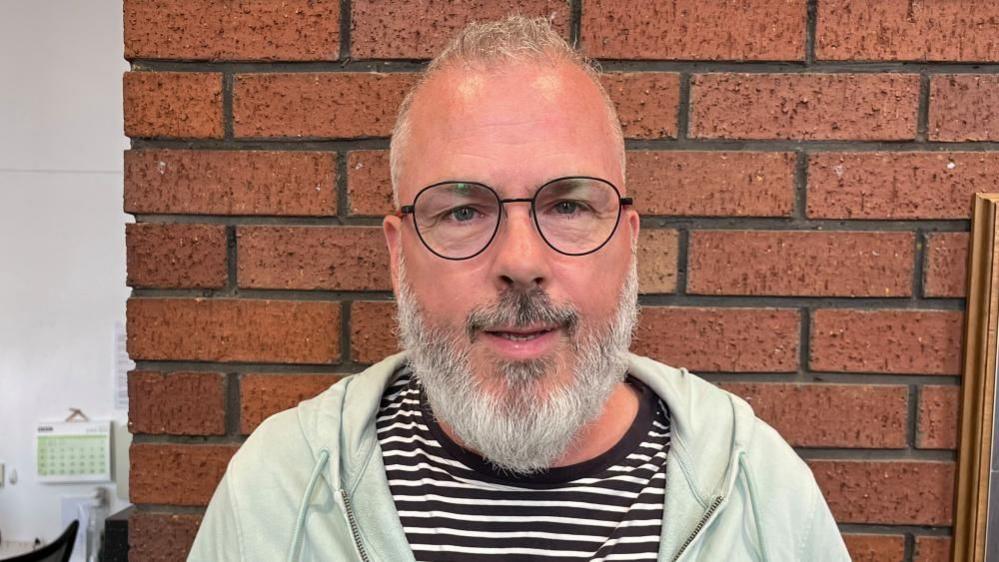 Graham Croucher with short white beard wearing a light green top in front of a brick wall