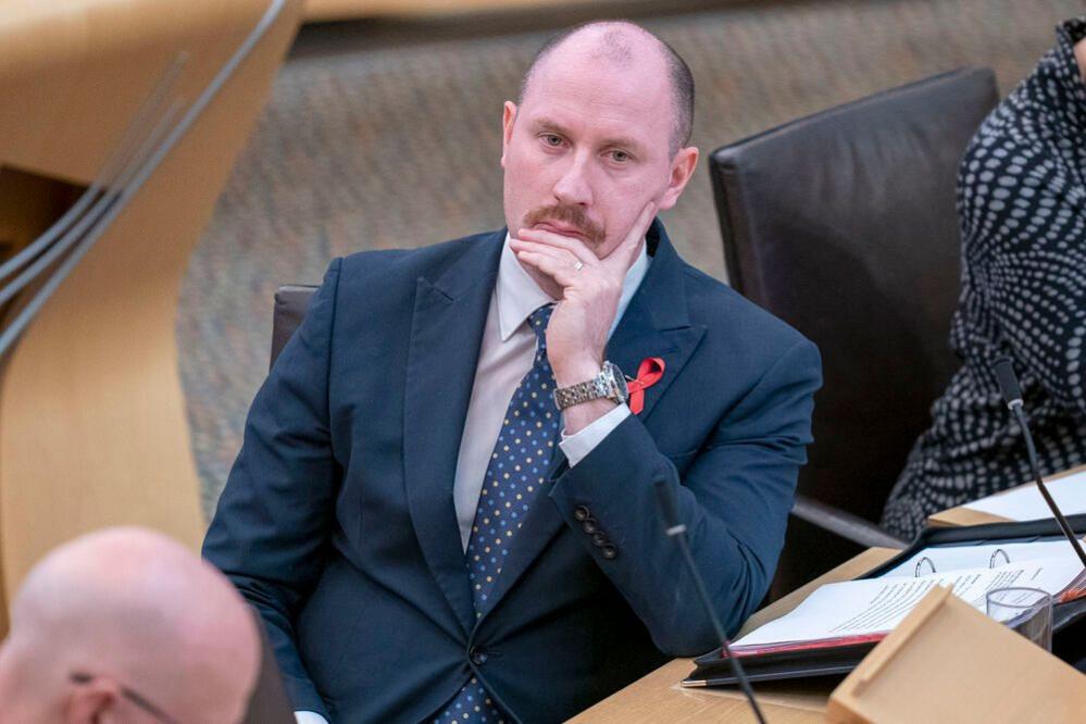 Neil Gray wears a dark suit and sit at a desk in the Scottish parliament chamber watching the first minister John Swinney speak.