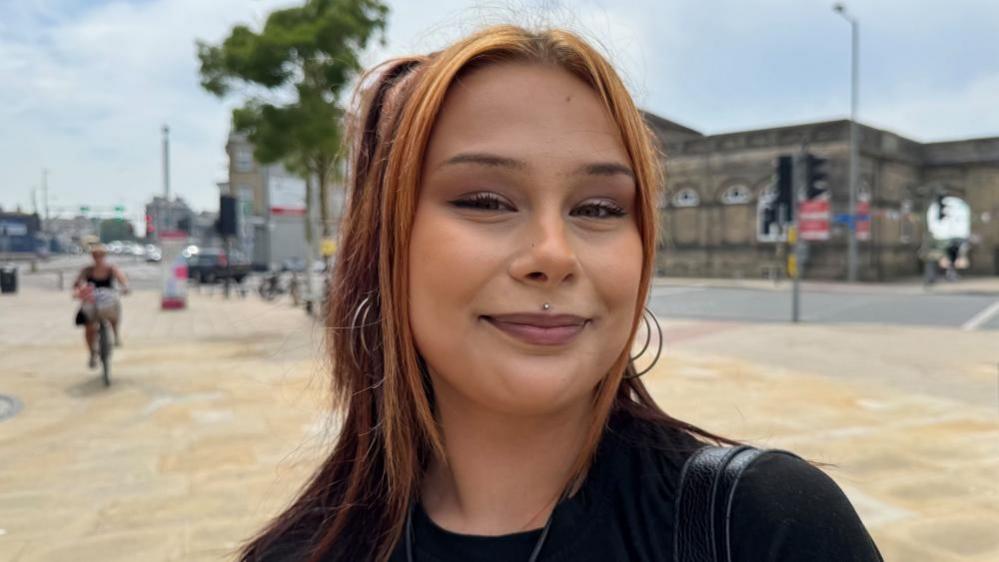 Poppy Stopforth has auburn hair, and is smiling. She has a piercing on her top lip; a stud, and she is pictured on the Station Square in Lowestoft, with Lowestoft Central railway station in the backdrop. A person riding a bicycle, in soft focus, is riding towards the camera, to the left of Poppy.