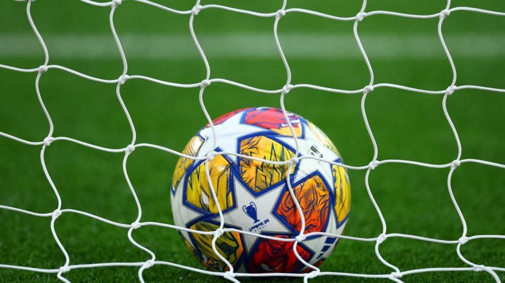 A white and orange football sits in a net on a green pitch. 