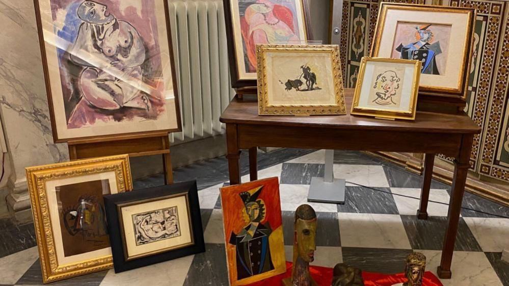 Copies of Picasso paintings are displayed on a table. Other artworks lean against an easel