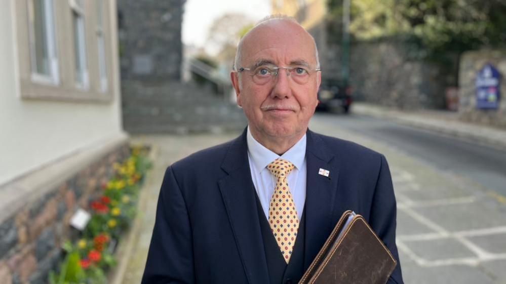 Man wearing glasses, a navy blazer and a yellow tie holding a brown folder. He is standing outside a grey building with flowers in the background