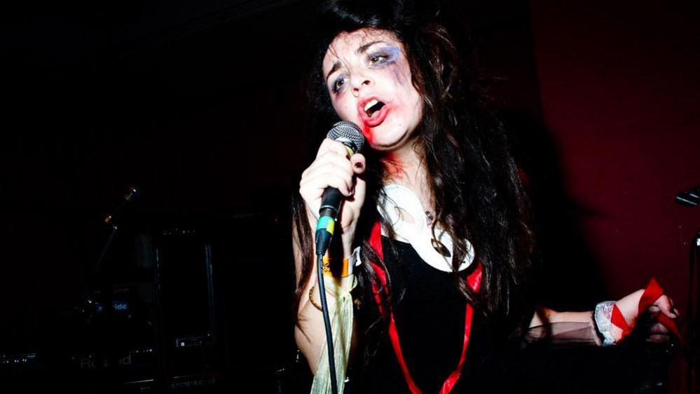 A woman singing into a microphone with a long red ribbon around her with hair that is dishevelled and make up that is smudged