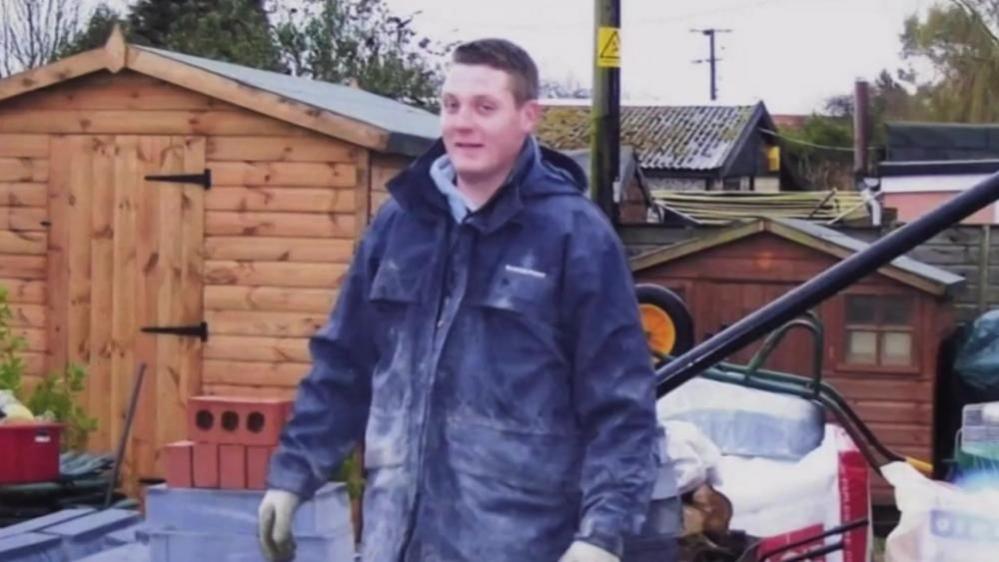 Terry McSpadden dressed in a blue coat, in the garden of a house where he had been working. In the background is a garden shed, a child's shed or wendy house, an upturned wheelbarrow and a drain pipe at a 45 degree angle. Behind Terry are more sheds and a telegraph pole with a yellow warning sign on it.