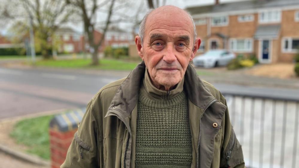 Nigel Costin, standing outside a row of homes, which are blurred in the background. He is wearing a green jumper, top, and coat, the coat is open. He is looking straight at the camera.