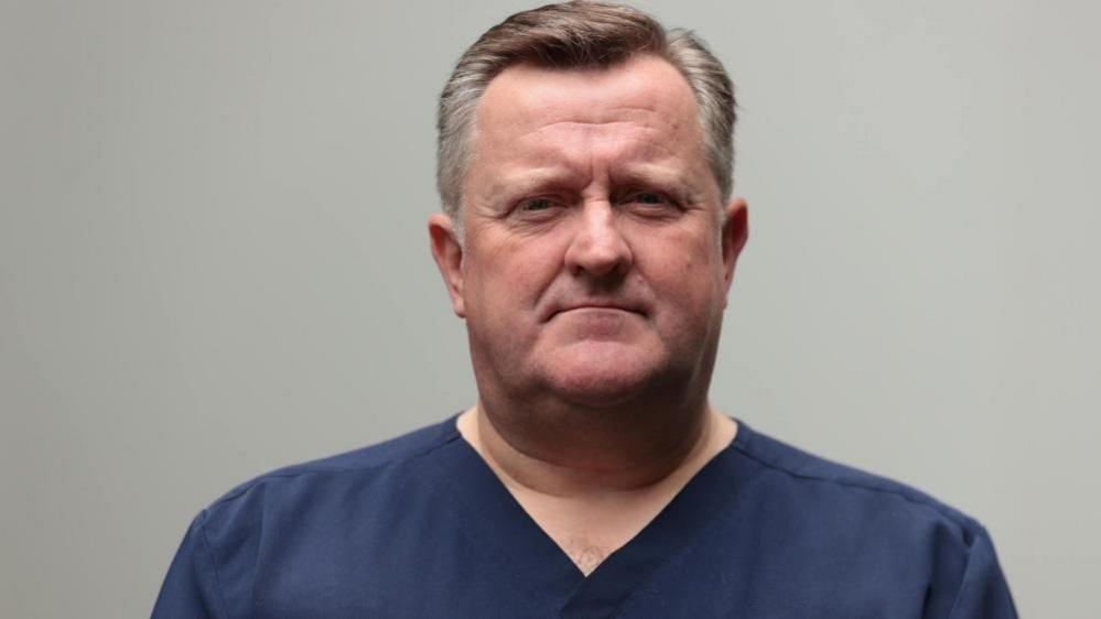 A man looks into the camera with a straight face. He has brown and white hair combed to the side. He is wearing dark blue medical scrubs.