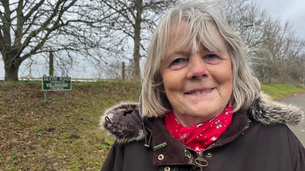 Penny Carpenter smiling at the camera, she is wearing a brown jacket and red scarf stood by a rural road sign and tree