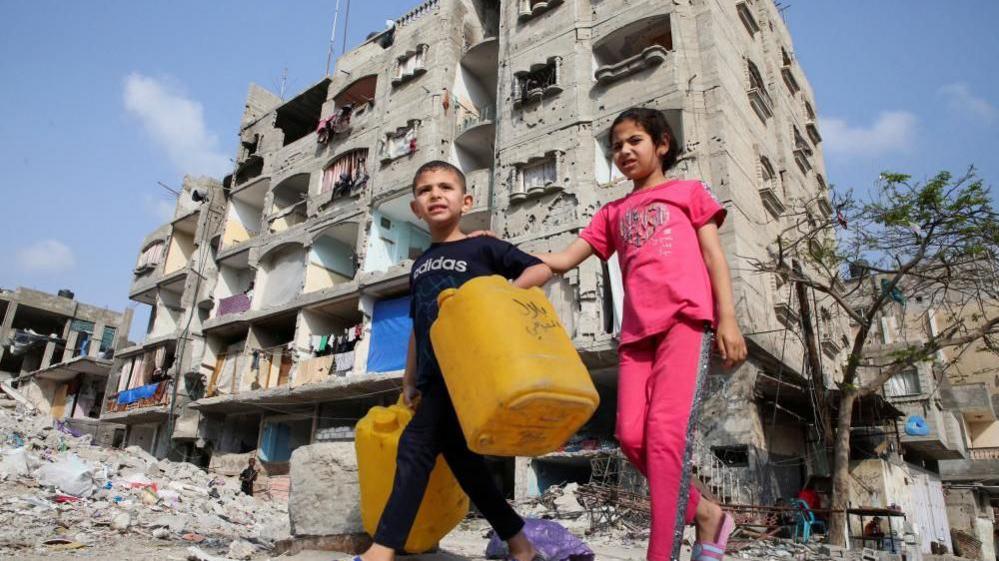 Two children carrying water butts walk in front of a bombed out building in Gaza