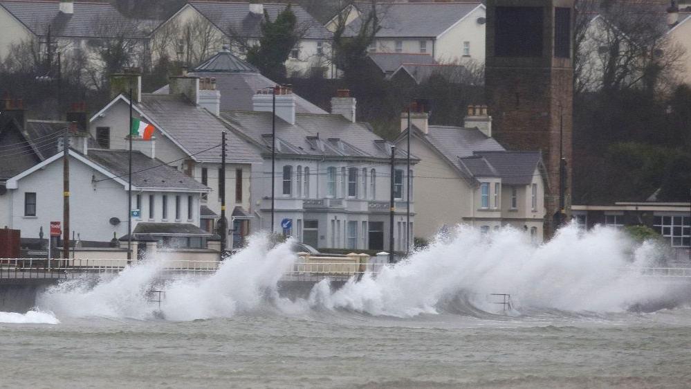 Waves hitting the sides of a coastal route with multiple buildings behind.