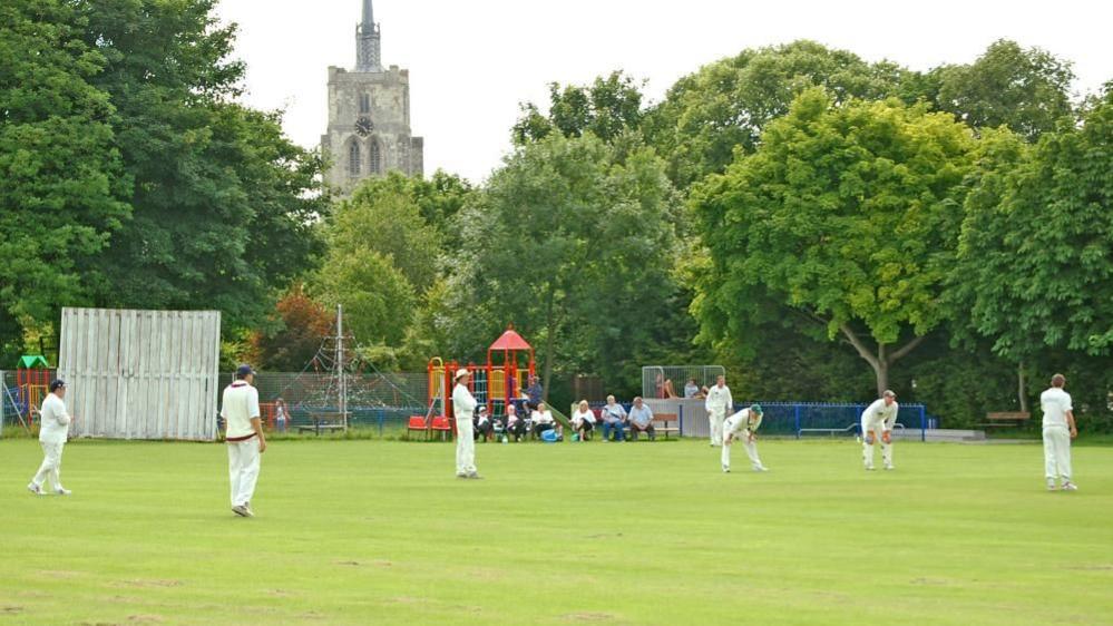 A village cricket match in Ashwell, Hertfordshire, on a summer's day in 2008. Cricketers in their whites are playing on a pitch in front of a church and are being watched by spectators.