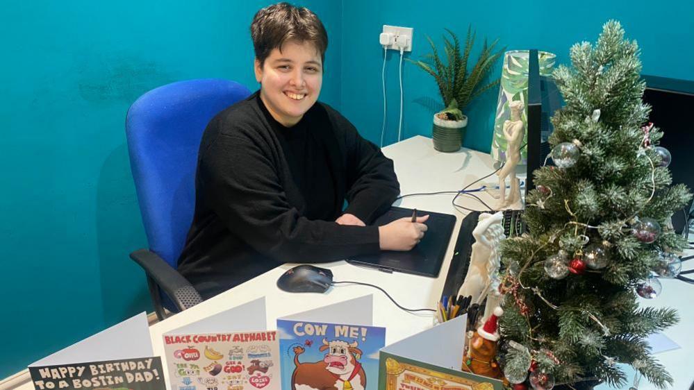 A lady sitting at a desk, with a computer in front of her. She is sitting on a blue chair and is wearing a black jumper. There is a small Christmas tree on the desk with lights and baubles, along with greetings cards