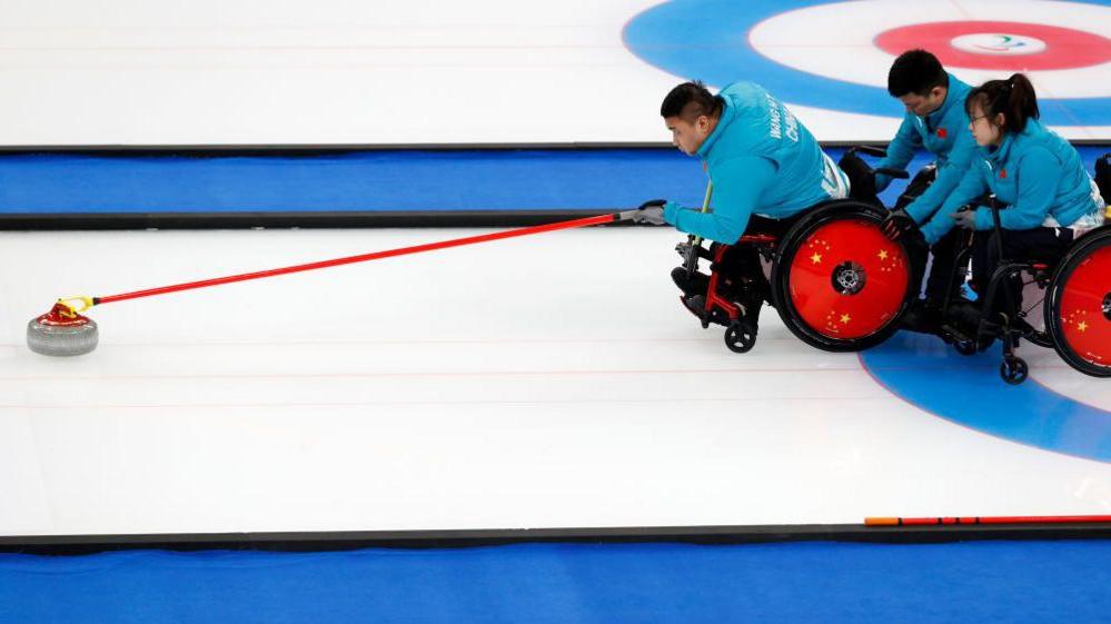 China deliver a stone in the wheelchair curling mixed team competition final at the Beijing Winter Paralympics. One player is holding a delivery stick with a curling stone at the end ready to play the stone while two players hold his chair for support