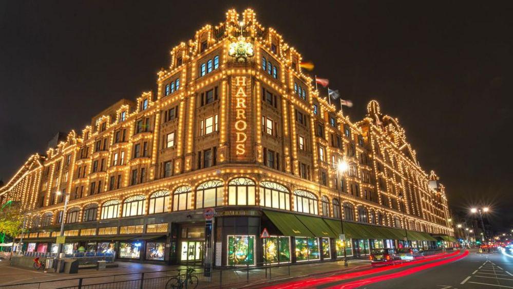 Picture of Harrods taken in the dark. It has seven floors and many windows, with white bulbs outlining the edges. The large shop windows on the ground floor have dark green awnings. 