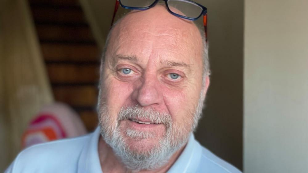 A man wearing a light blue top stands in his hallway. The walls are pale and wooden stairs can be seen behind him.