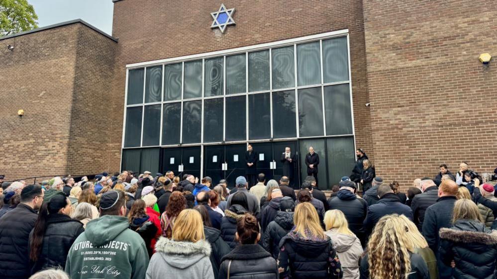 A large crowd of people stand with their heads bowed in front of a large brick building with a Star of David on the front. People are stood on a stage in front of them.