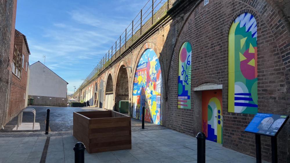 A walkway with brightly coloured red brick arches on the right. There is an open wooden box in the middle of the path, that could be for plants and silver bike racks on the left. The sky is blue in the background. 