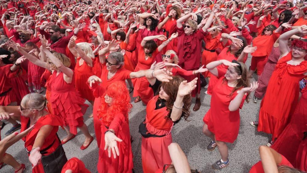 A group of people  in red dresses dance, holding their arms in the air.