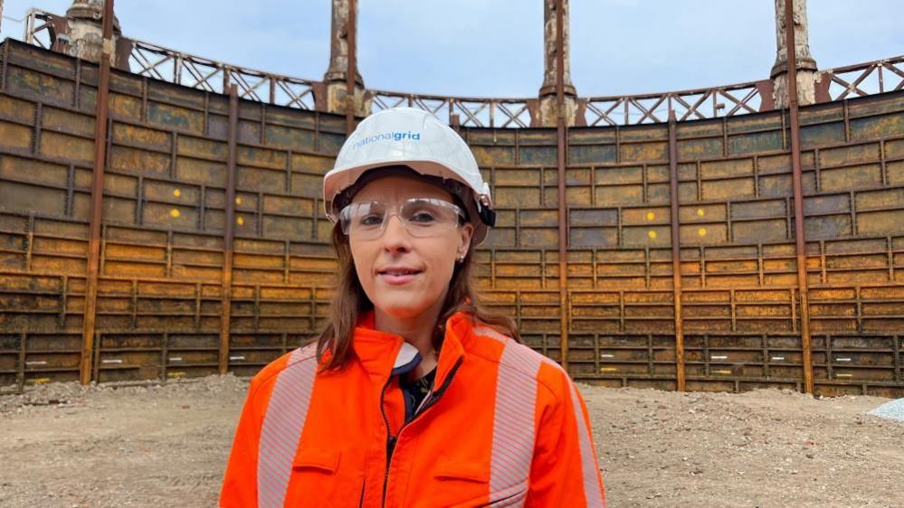 Nadia McInnes is wearing a white hard hat and orange high visibility jacket. She has brown hair, which is at shoulder length, and she is wearing safety glasses. She is standing within the tank are of the Great Yarmouth gasometer, which has had the lid removed, and exposed the inside structure of the rusting cast iron tank which shows ribs and vertical rails.