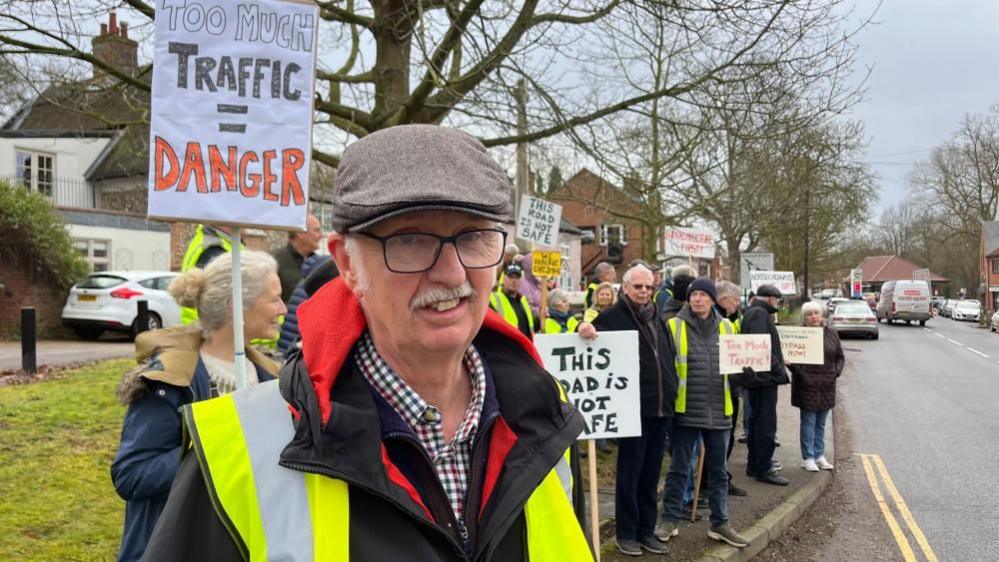 Bill Musson is standing near a group of protestors, with placards, reading: "Too much traffic = danger"; and: "This road is not safe", with others in the distance. Mr Musson is wearing a blue, white and red checked shirt, a black jacket with red inside lining and a high visibility yellow vest. He has a grey moustache, wears black rimmed glasses and is wearing a tweed flat cap.