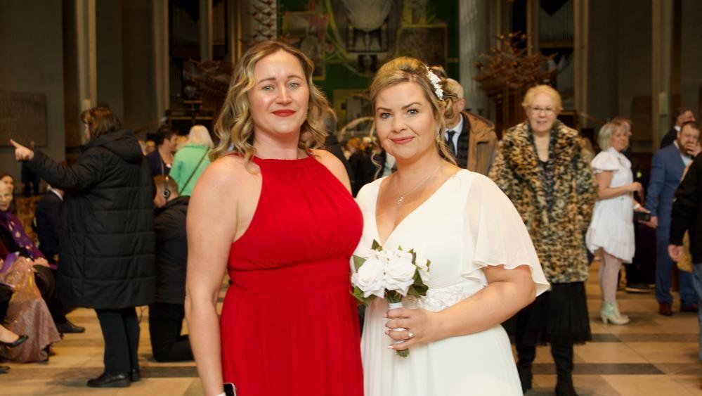 Two women standing in a church. One is wearing a long red dress and one is in a white wedding dress, holding a bouquet of flowers 