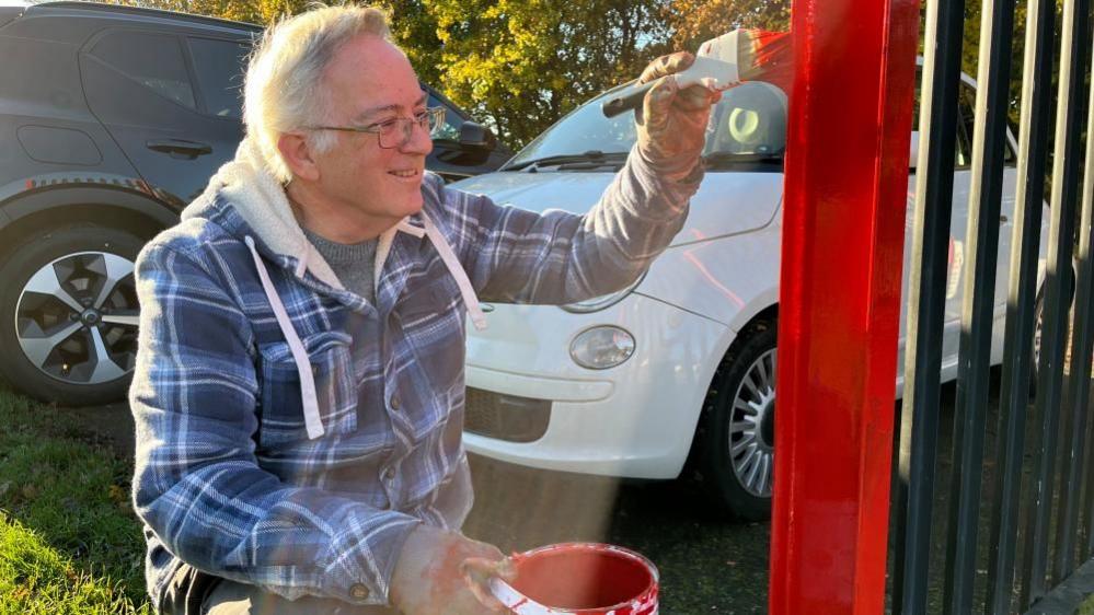 A man with grey hair and glass painting a fence with red paint. There is a black car and white car behind him.