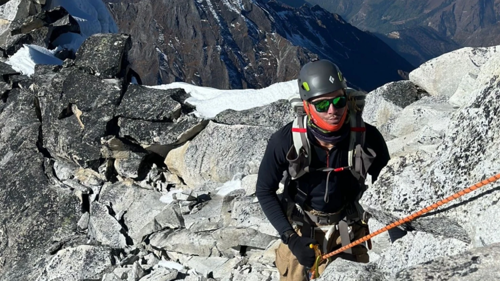 Rhys Fitzgerald climbing in mountains. He is wearing climbing gear and holding a rope on a rocky section, with mountains and some snow behind him