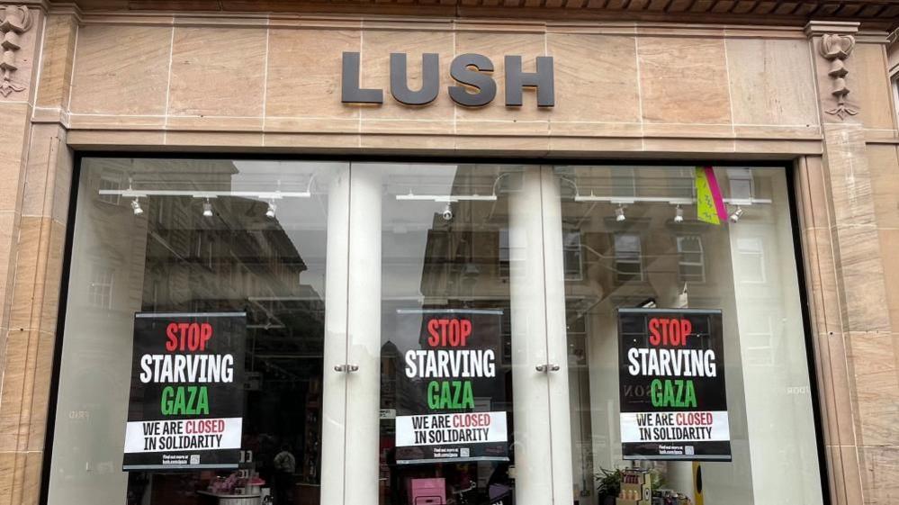 The shopfront of a Lush store with three posters in each window which reads: Stop starving Gaza, we are closed in solidarity. The lights inside are off