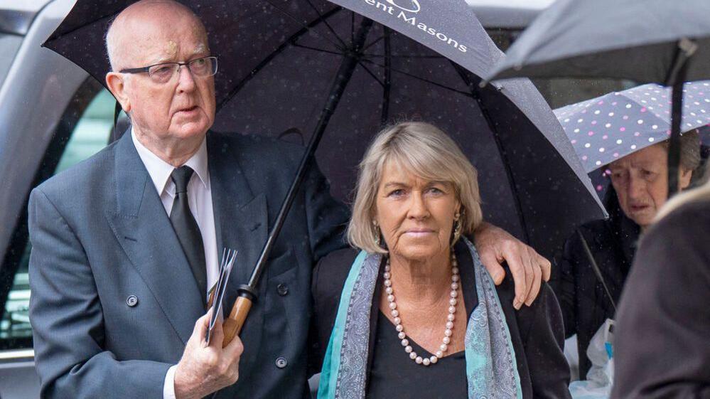 A man in a dark suit holds an umbrella over Rose Riley who wears dark clothes and looks at the camera