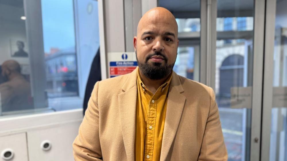 Daniel Arneaud in a yellow shirt and beige jacket looking at the camera.