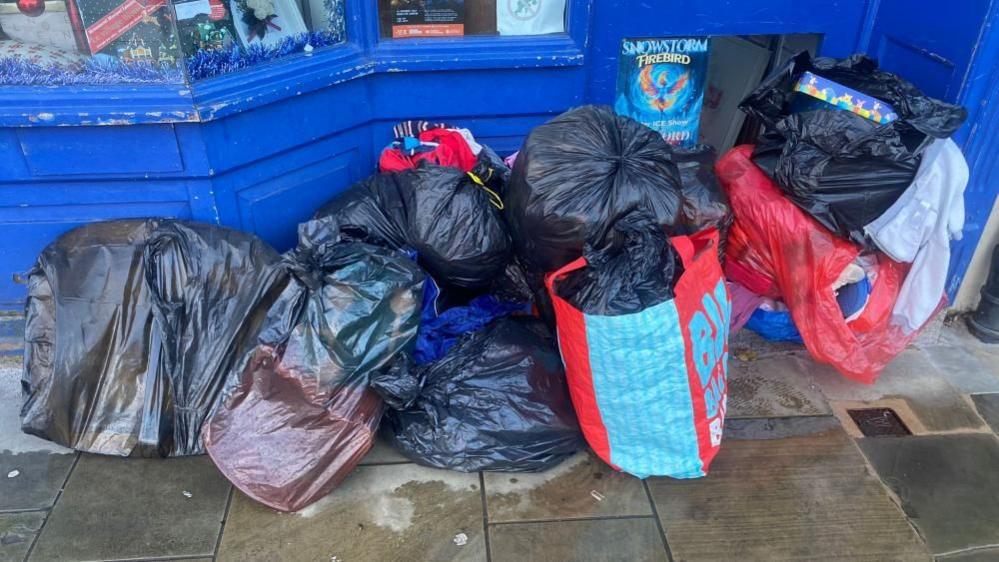 A pile of full black bin bags sit outside a shop door.