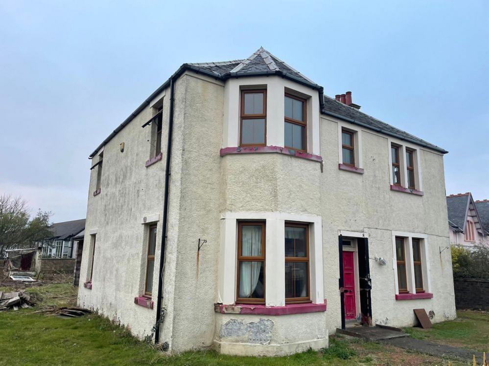 An abandoned and detached white house with a black roof, red door and brown window panes sits in a state of disrepair on grass.