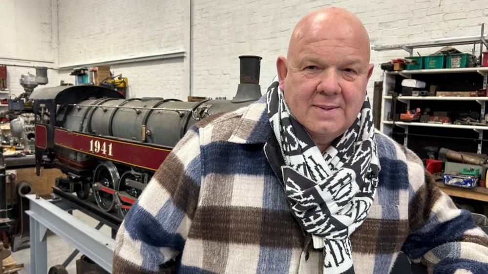 Joe Nemeth standing in front of a miniature steam engine with 1941 written on the side. He is a bald man, wearing a white jacket with blue and brown cross hatching, and a black and white scarf.