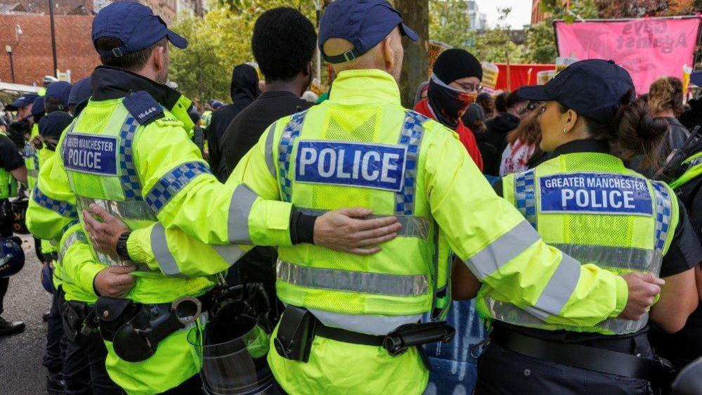 Greater Manchester Police form a barrier during the riots with hands around their shoulders. They are wearing neon yellow jackets reading Greater Manchester Police on a block of blue on the jacket. 